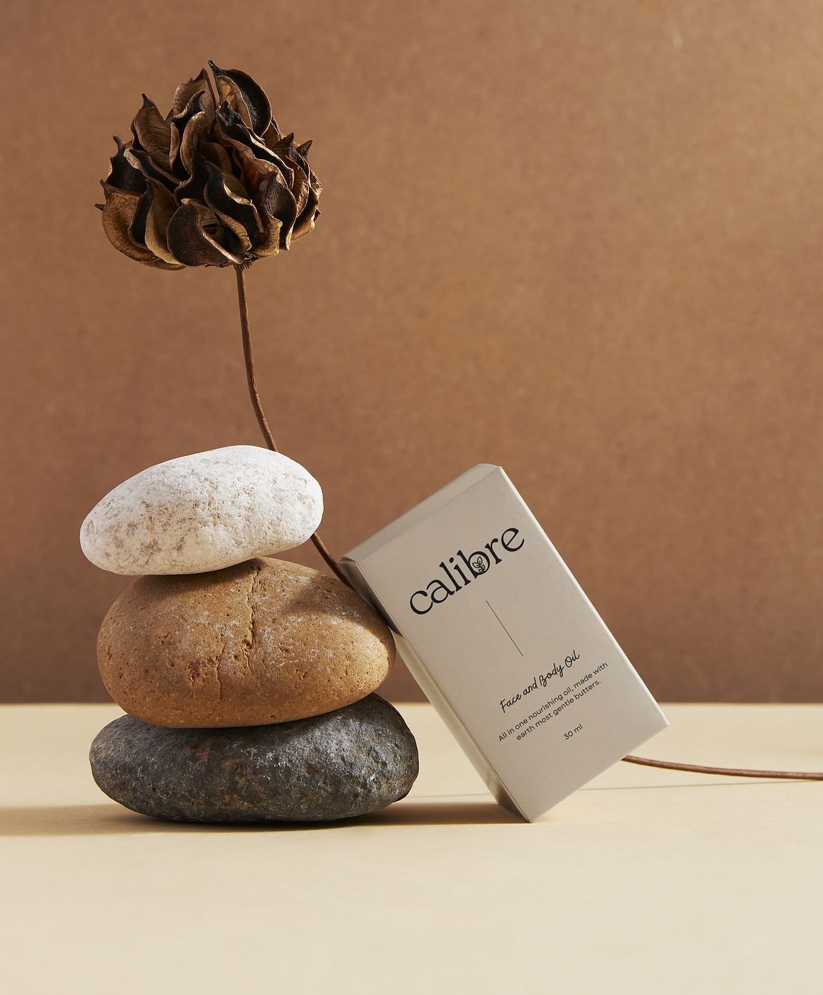 Calibre face and body oil box leaning on stacked stones with a dried flower