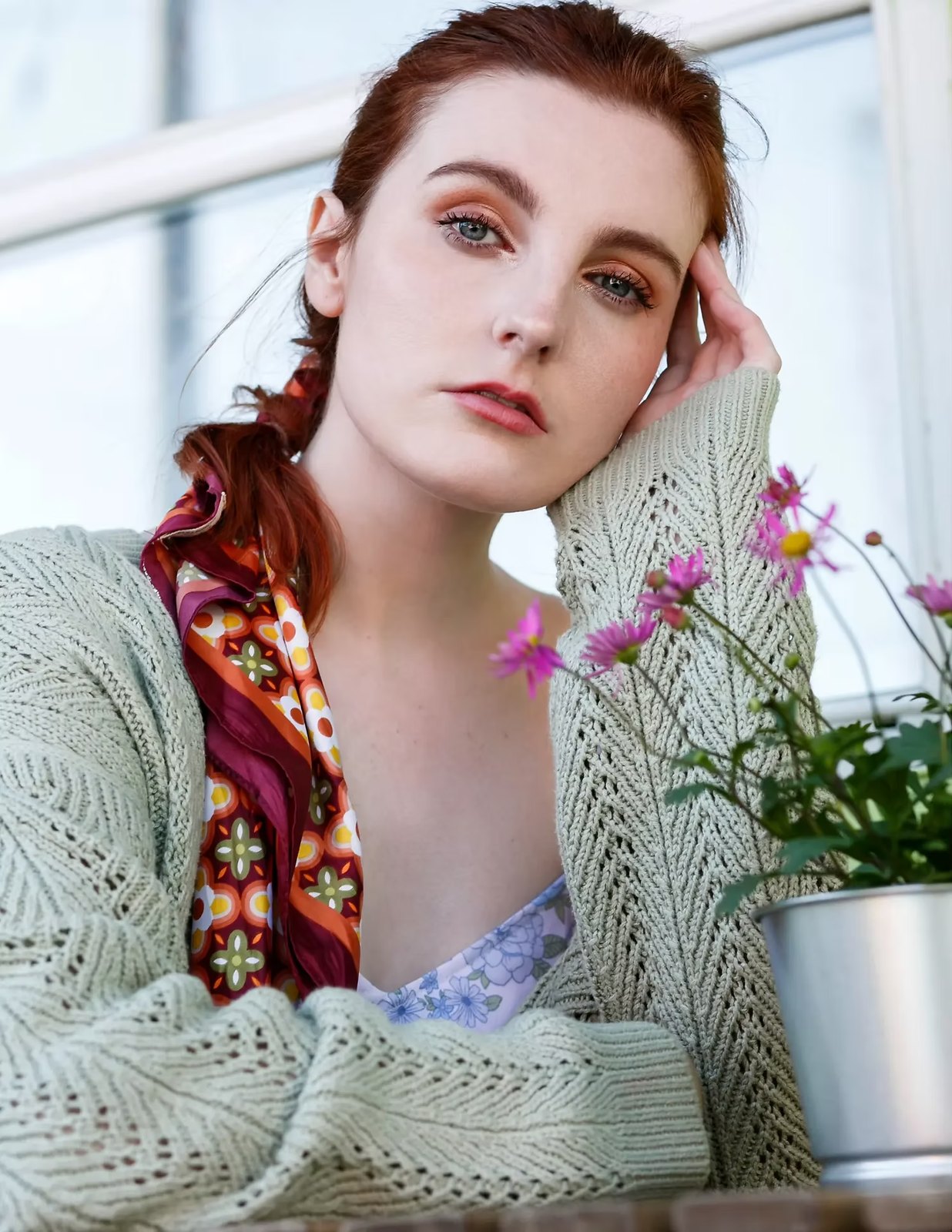 Red-haired model in a mint cardigan resting her head on her hand beside pink flowers