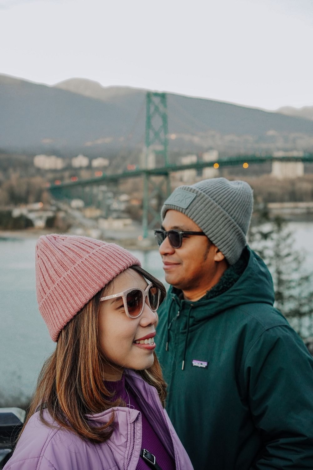 Couple in winter beanies smiling in front of the Lions Gate Bridge
